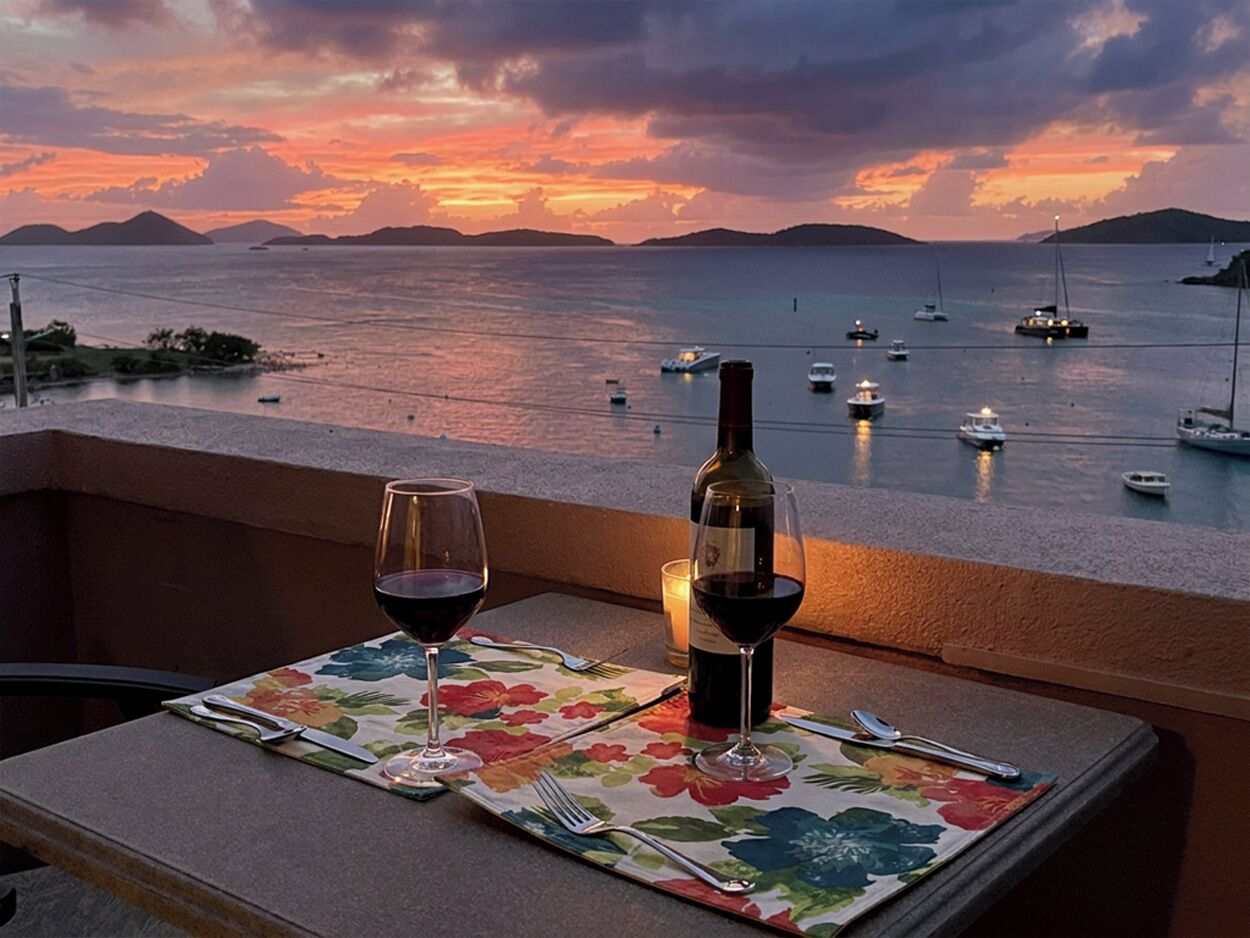 A table set with a bottle of wine and glasses, overlooking a calm sea with boats and a sunset.