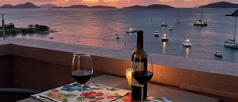 A table set with a bottle of wine and glasses, overlooking a calm sea with boats and a sunset.