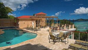 A pool area with lounge chairs, a building with a red roof, and a view of the sea. - Stunning Penthouse at Grande Bay - Breathtaking Views and Luxury Living! (St John)