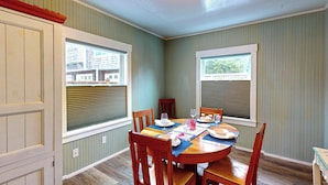A dining area with a round table set for four, flanked by wooden chairs, a window with blinds, and a view of buildings outside. - Cozy Cottage at Seaside Beach (Seaside)