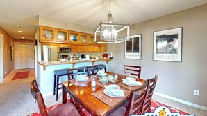 Uma sala de jantar com uma mesa de madeira posta para seis lugares, cozinha com armários de madeira, micro-ondas e geladeira.