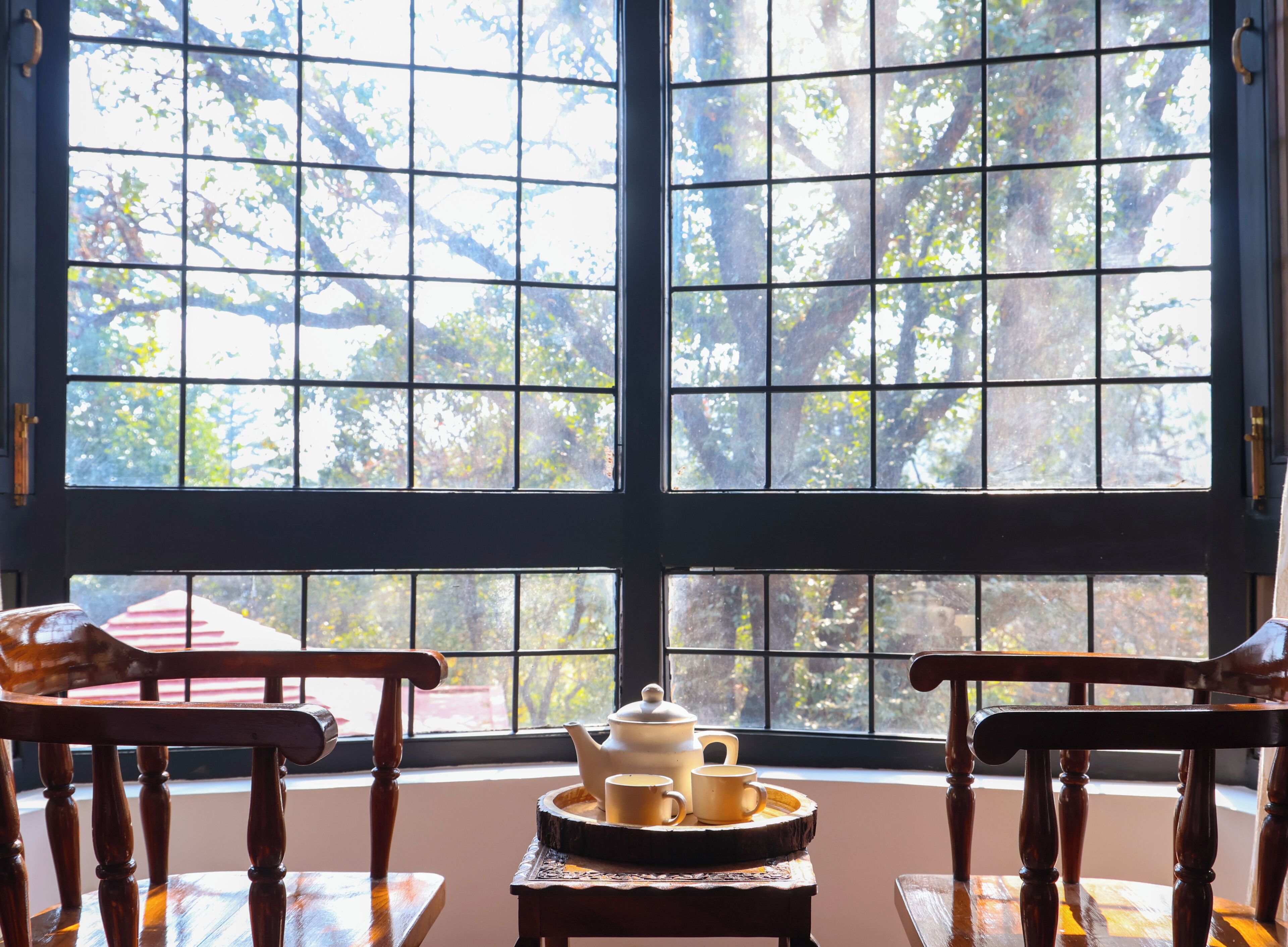A room with a large window, two wooden chairs, a small table with a teapot and cups, and a view of trees outside.