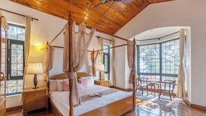 A four-poster bed with a canopy, wooden flooring, a nightstand, a bedside table with a lamp, and a view of the outdoors through large windows. - Alaya Stays Thistle House 4 BHK Hideaway (Bhimtal)