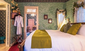Una camera da letto con un letto, una sedia di legno, un accappatoio e una porta che dà su un'altra stanza.