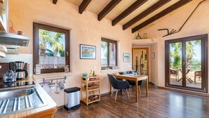 Una cocina con un lavamanos, estufa y estanterías de madera; una zona de comedor con mesa y sillas; y ventanas amplias con vistas a palmeras.