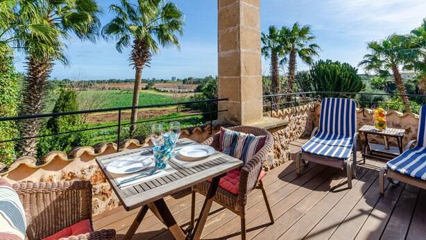 Una terraza con mobiliario de mimbre, una mesa dispuesta para comer y vistas a palmeras y campos.