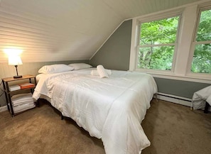 Un dormitorio con cama, una mesita de noche con lámpara, una ventana con vistas a árboles y un radiador.