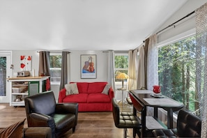 Uma sala de estar com um sofá vermelho, uma mesa de jantar com cadeiras, uma estante de livros e uma janela com vista para árvores.