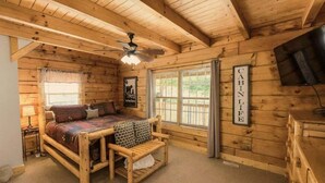 Habitación tipo cabaña con cama de madera, un banco, ventilador de techo, televisión y ventana con cortinas.