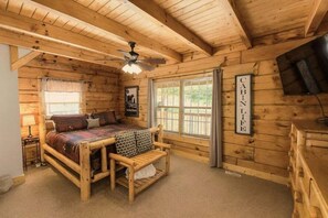 Habitación tipo cabaña con cama de madera, un banco, ventilador de techo, televisión y ventana con cortinas.