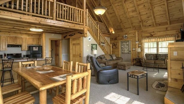 Una cabaña espaciosa con interior de madera, un comedor, una sala de estar con sofá y sillones, y una cocina con electrodomésticos modernos.