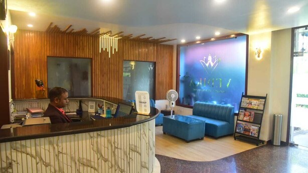 A reception area with a wooden reception desk, a blue sofa, a fan, and a wall with a logo. - HOTEL VEDIKA  (Ranchi)