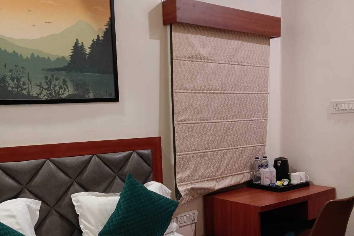 A hotel room with a bed, a desk, a chair, a painting, and a window with a view of a mountain and trees.