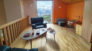 A room with a wooden ceiling, a black leather chair, a small table with chess pieces, a radiator, and a view of a house through a window. - Sea View House for 9 - Beach Just Around the Corner & Family Comfort (Sarzeau)
