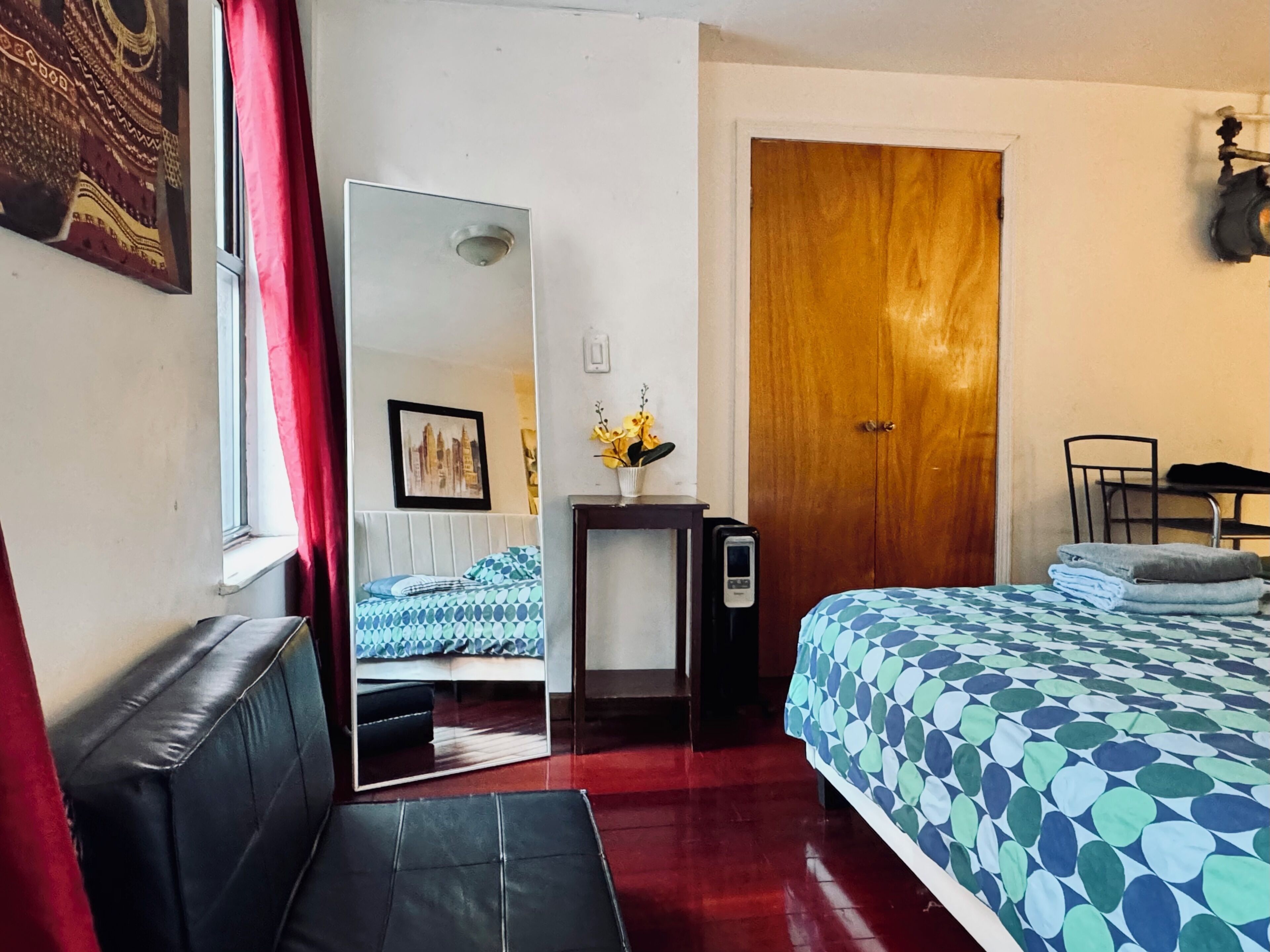 A hotel room with a bed, a desk, a chair, a mirror, a door, and a window with red curtains.