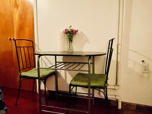 A small dining table with two chairs, a vase with flowers, and a white wall with a door and electrical outlet.