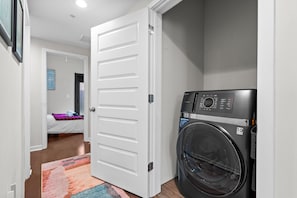 Ein Waschraum mit einer Waschmaschine und einem Trockner, ein Flur, der zu einem Schlafzimmer mit einem Bett und einem farbenfrohen Teppich führt.