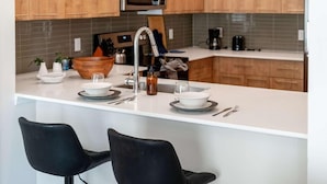 A modern kitchen with wooden cabinets, a white countertop, a built-in microwave, and two black bar stools. - Riviere 2BR Apartment Pet Friendly Oasis Sleeps 6 Close to Epic Universe Pool & Gym (Orlando)