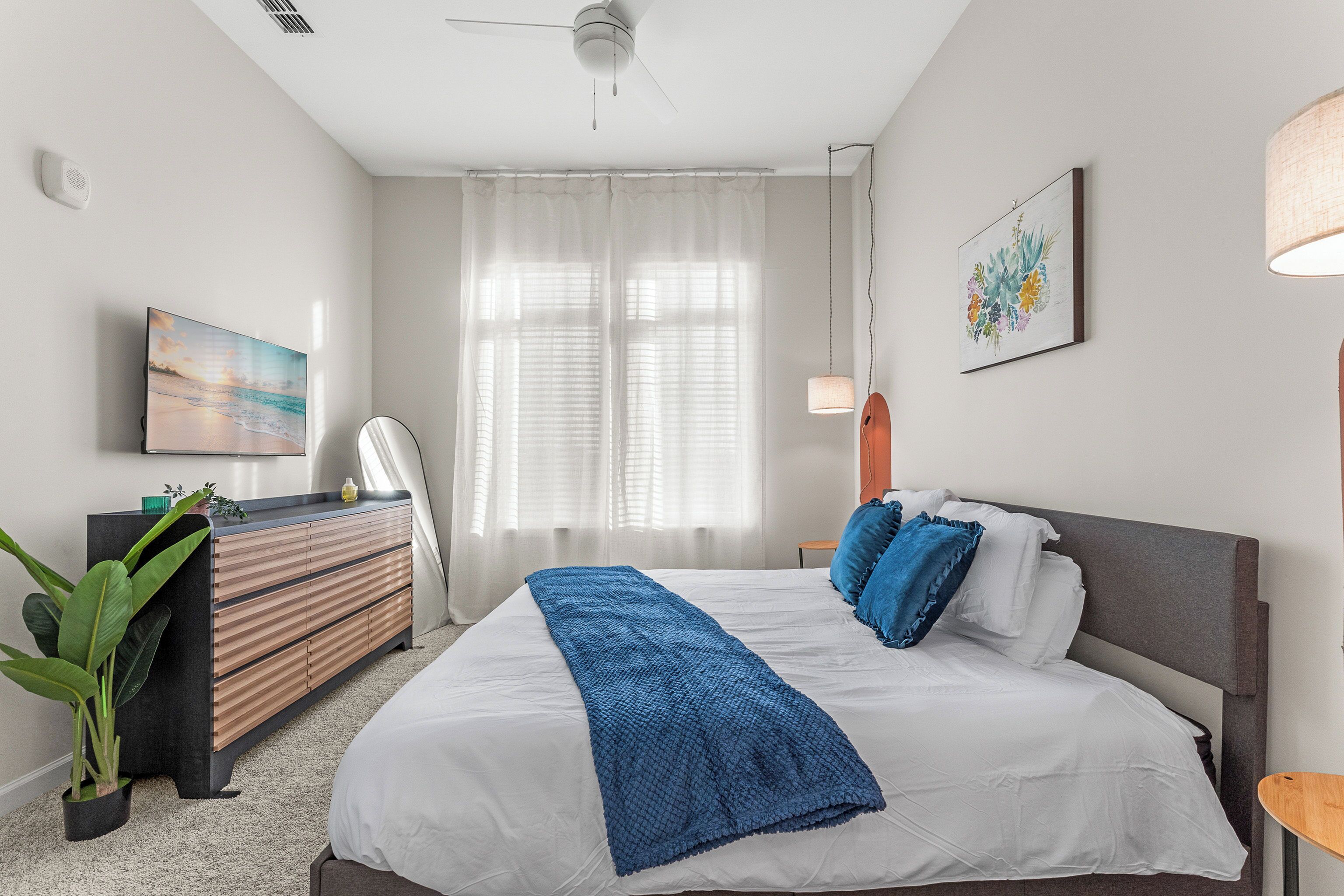 A bedroom with a bed, a dresser, a plant, a ceiling fan, and a window with curtains.