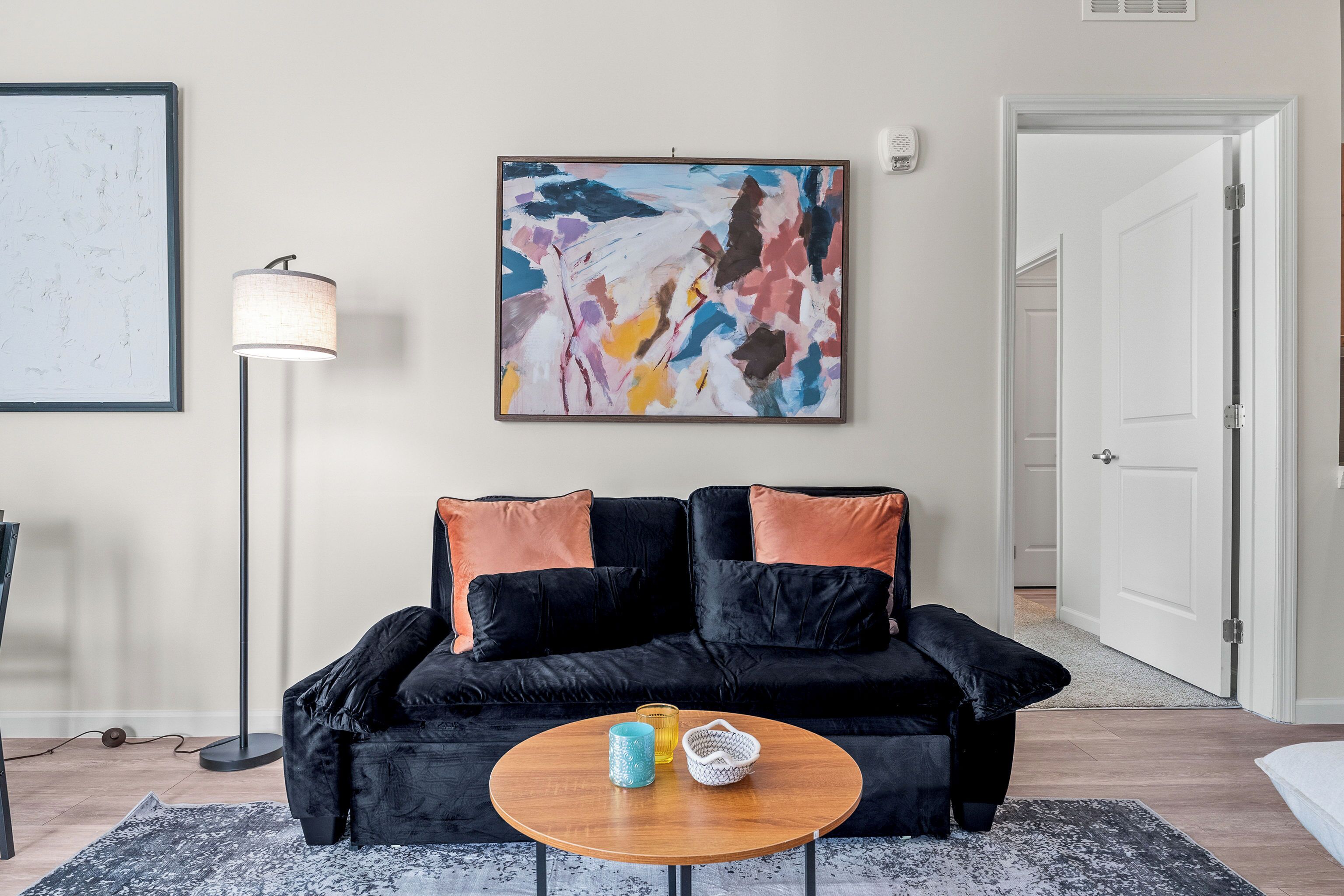 Un salon avec un canapé noir, une table basse ronde, un lampadaire et un tableau abstrait aux couleurs vives.