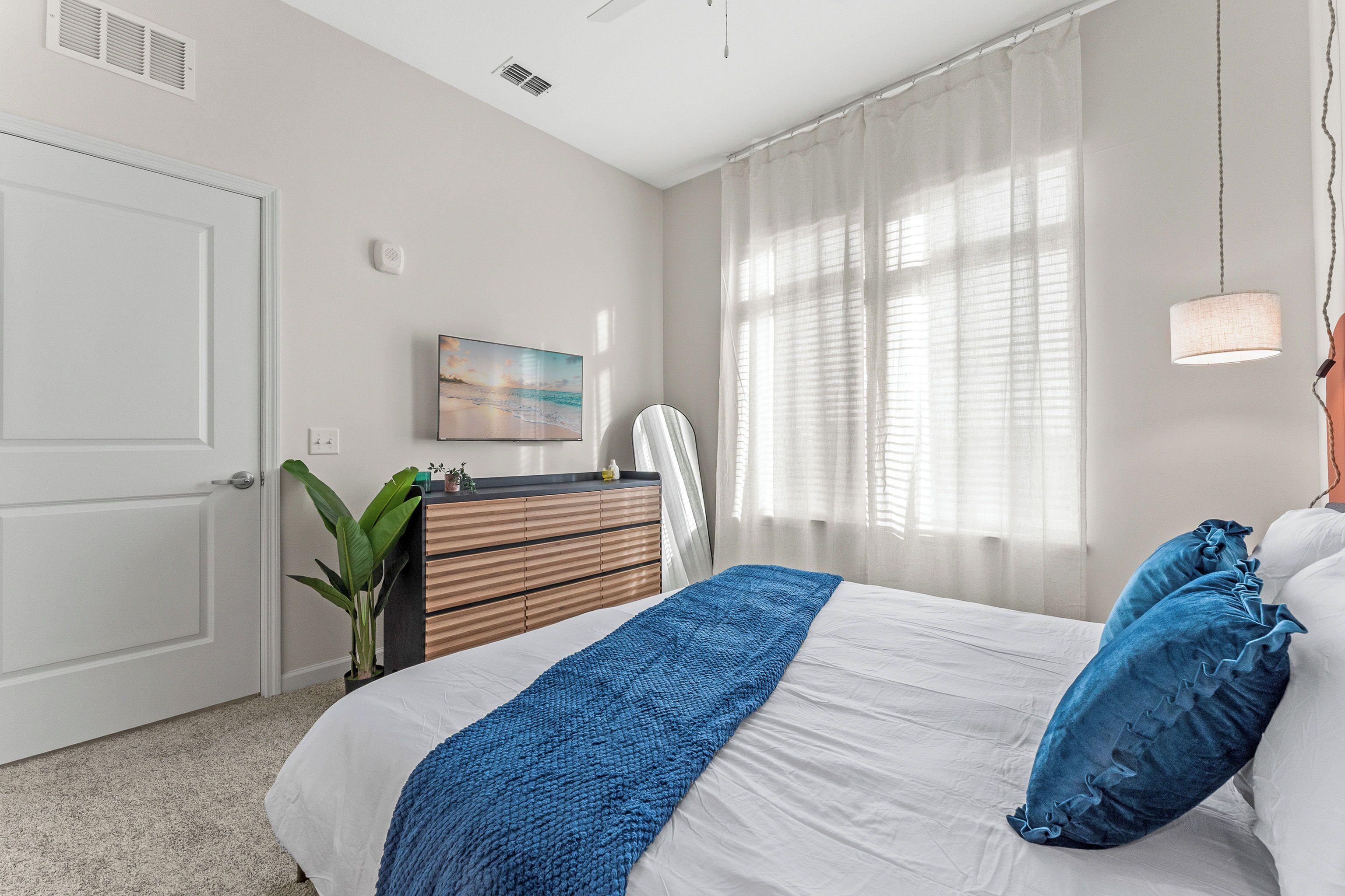A bedroom with a bed, a dresser, a plant, a painting, and a window with curtains.