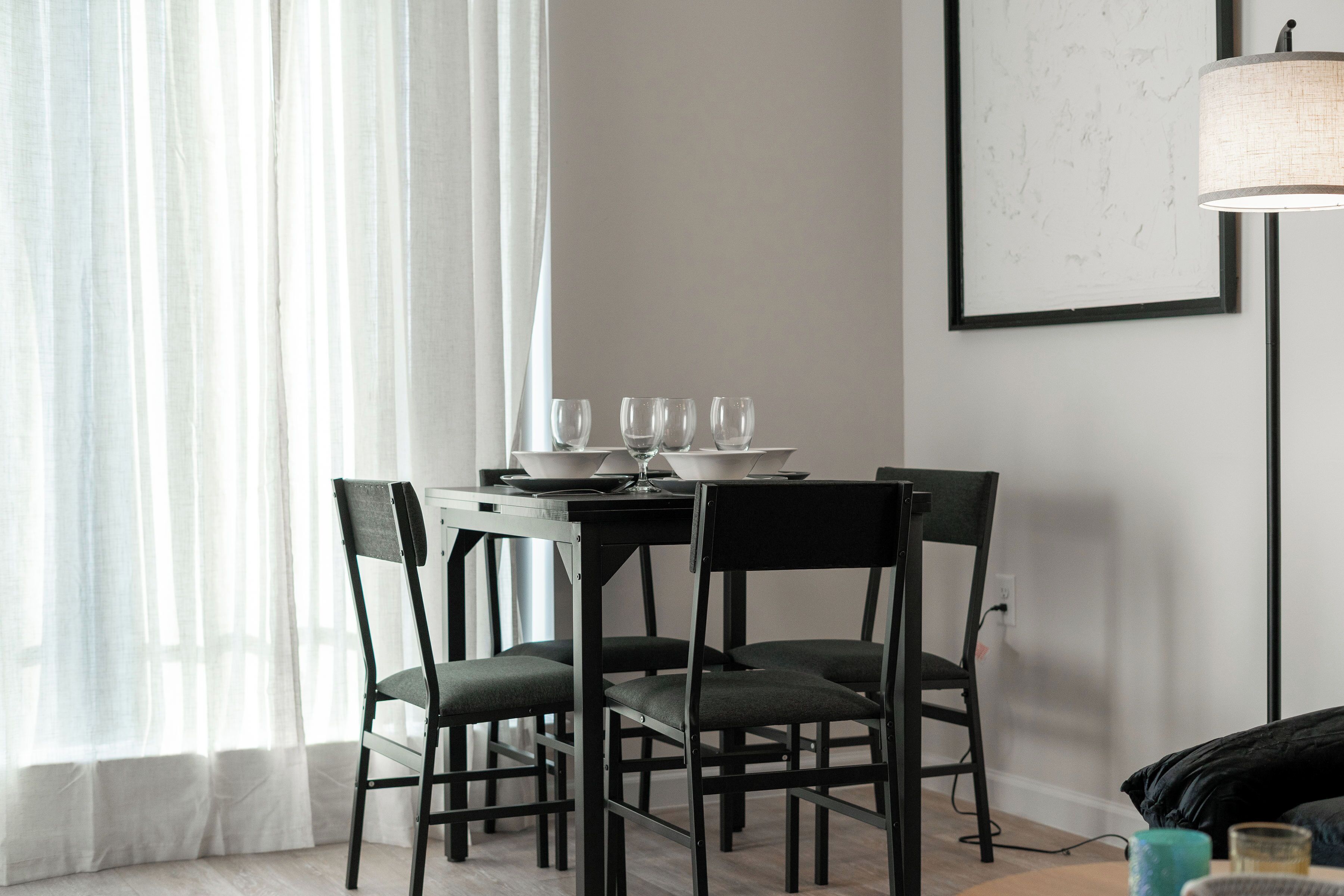 Uma sala de jantar com uma mesa posta para quatro pessoas, cadeiras, um abajur e um quadro na parede.