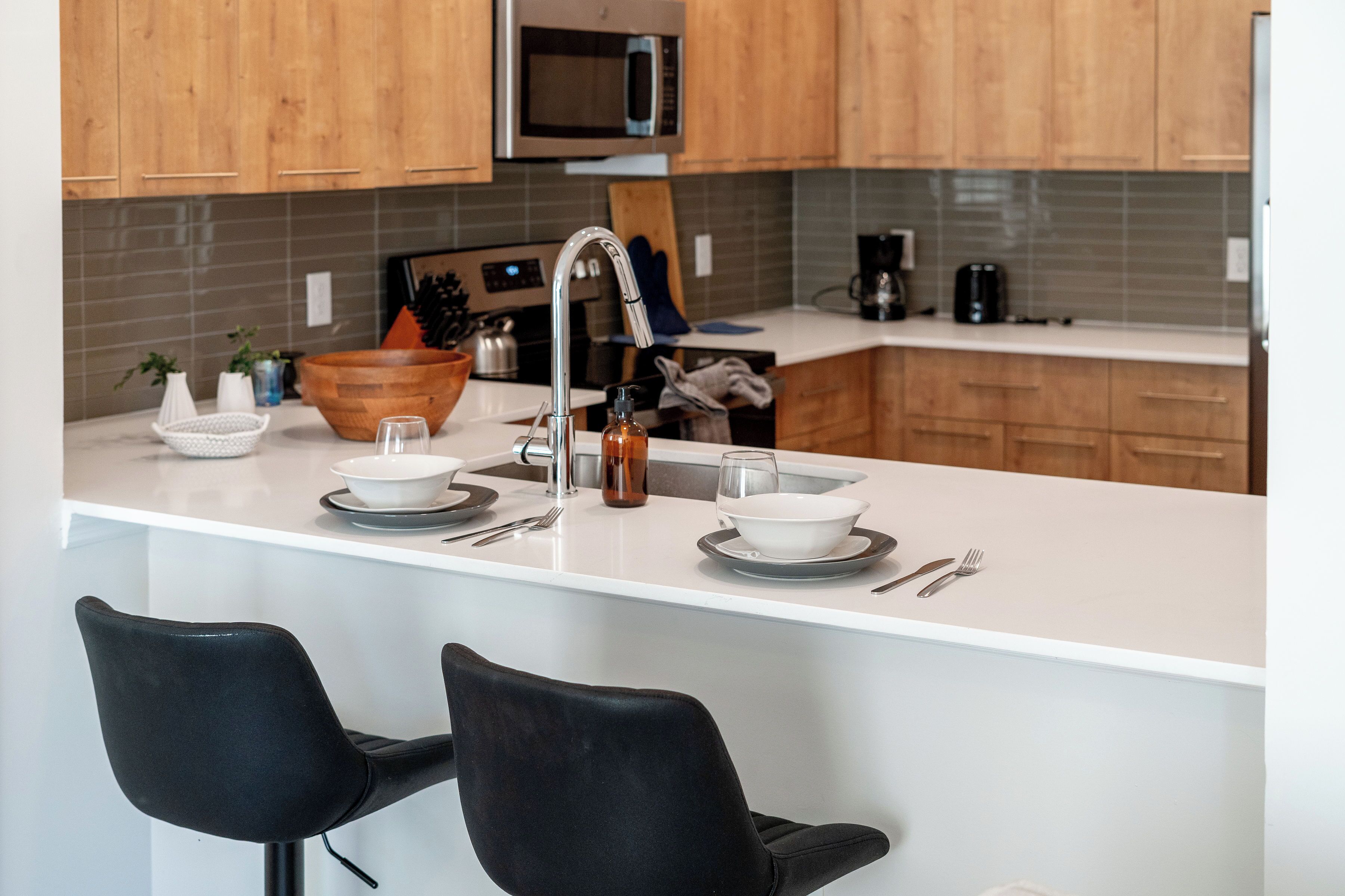 A modern kitchen with wooden cabinets, a stainless steel microwave, a white countertop, and two black chairs.
