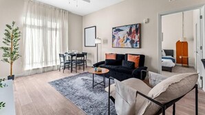 A modern living room with a black sofa, a dining table with chairs, a ceiling fan, and a large abstract painting. - Riviere 2BR Apartment Pet Friendly Oasis Sleeps 6 Close to Epic Universe Pool & Gym (Orlando)