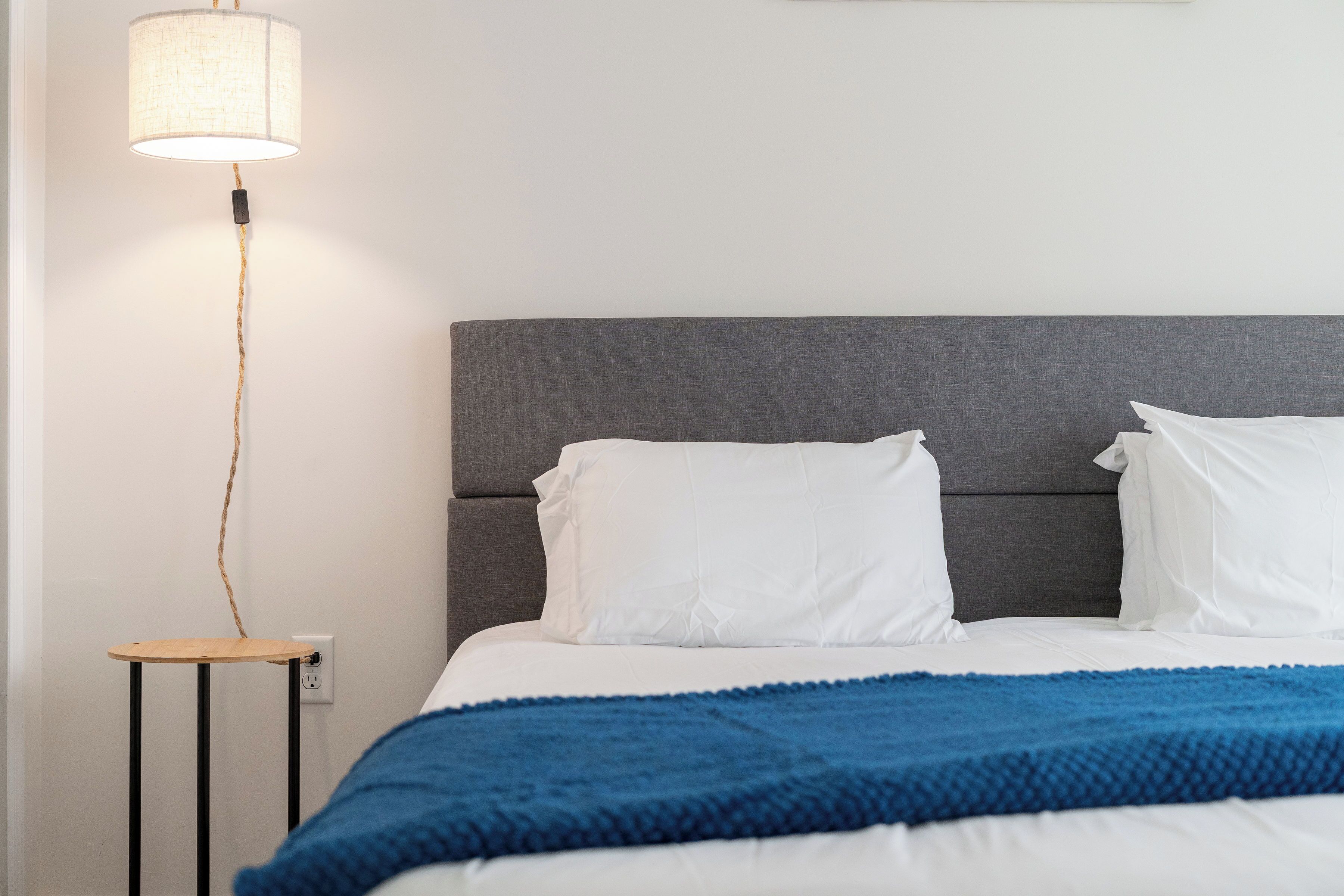 Ein Bett mit weißer Bettwäsche und einer blauen Decke, ein grauer Kopfteil, ein Nachttisch mit Lampe und eine Wandsteckdose.