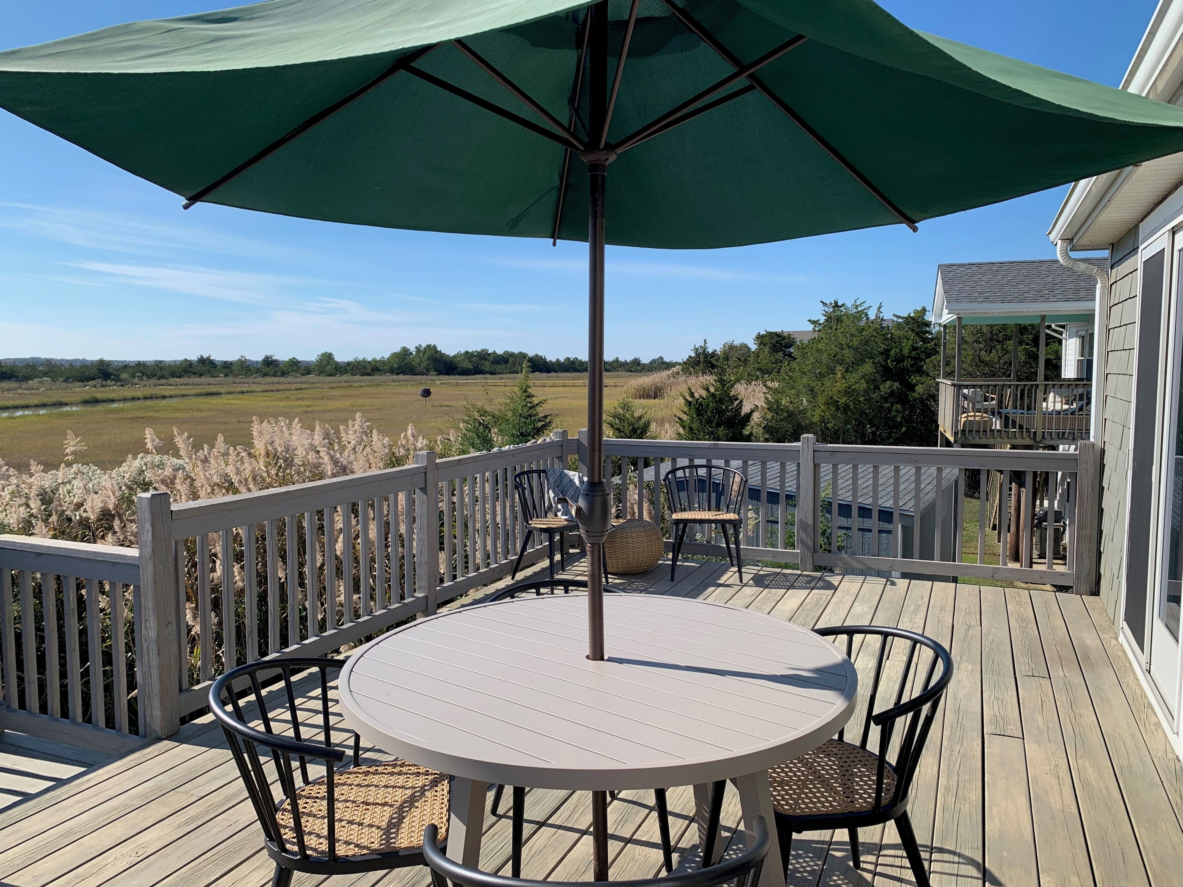 A wooden deck with a round table and chairs, a green umbrella, and a view of a field and trees.