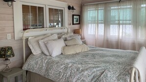 Un dormitori amb llit, una nitòria, una làmpada, una finestra amb cortines i una lluminària penjada a la paret.