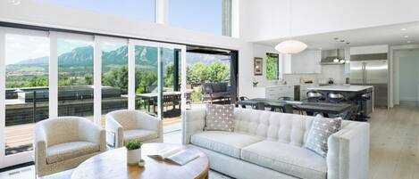 Ein großzügiger Wohnbereich mit einem großen Fenster und Blick auf eine Terrasse, ein Sofa, ein Couchtisch und eine Küche im Hintergrund.