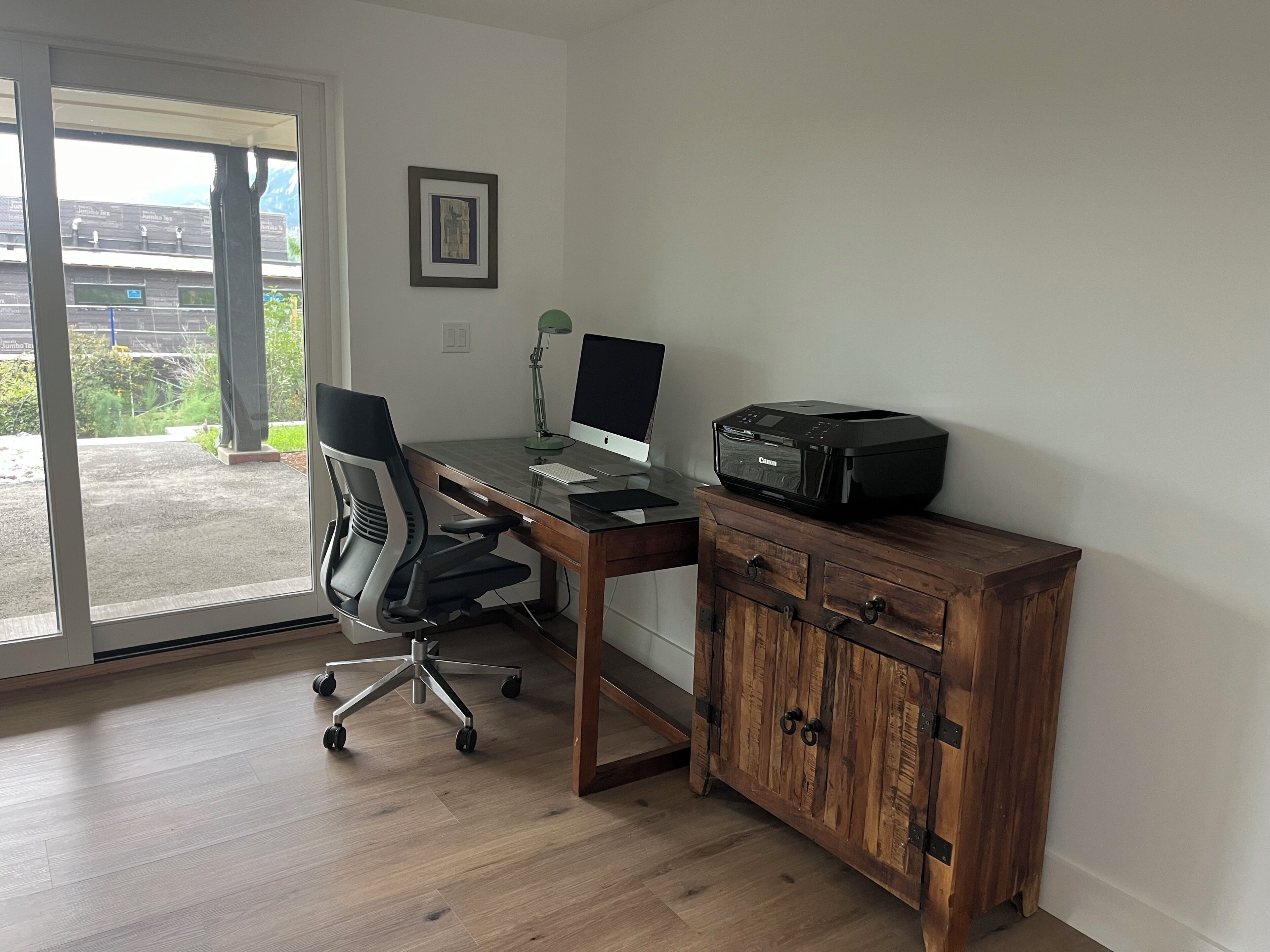 Una oficina en casa con escritorio, silla, computadora, impresora y vistas a través de una puerta de vidrio.