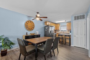 A kitchen with wooden cabinets, a large clock, a ceiling fan, and a dining area with a wooden table and wicker chairs. - Cozy 2 Bedroom with Ocean Views (Carolina Beach)