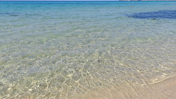 Klares, flaches Wasser, ein Sandstrand und in der Ferne felsige Felsen.