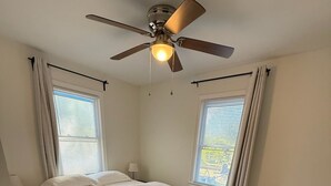 Ein Schlafzimmer mit einem Bett, Deckenventilator, zwei Vorhängen versehene Fenster, einem Teppich und einem Nachttisch.