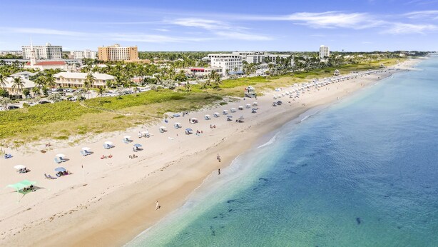 A beach with rows of sun loungers and umbrellas, a clear blue ocean, and buildings in the background. - Delray Breezes !NEW! WALK to BEACH & THE AVE! (Delray Beach)