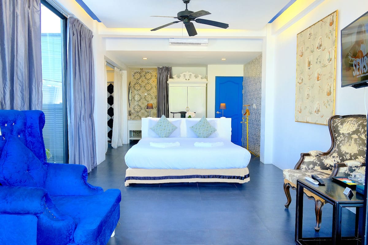 A hotel room with a large bed, a blue armchair, a small table, a ceiling fan, and a TV.