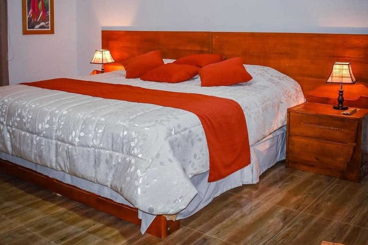 Habitación de hotel con cabecera de madera, cama grande con ropa de cama blanca y naranja, dos lámparas de noche y un cuadro en la pared.