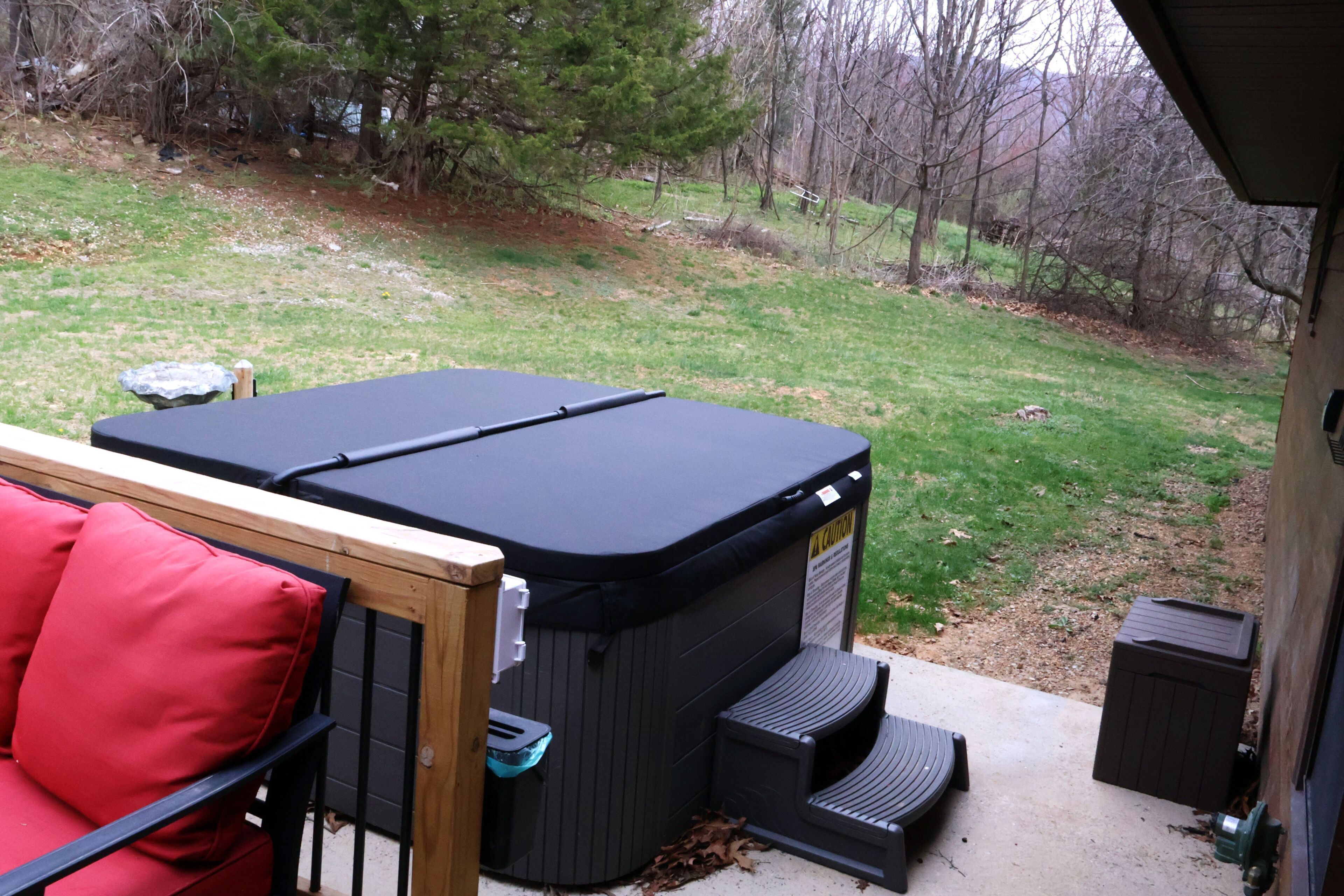 A hot tub with a wooden deck and red cushions, a concrete patio, and a grassy yard with trees.
