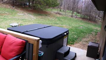 A hot tub with a wooden deck and red cushions, a concrete patio, and a grassy yard with trees.