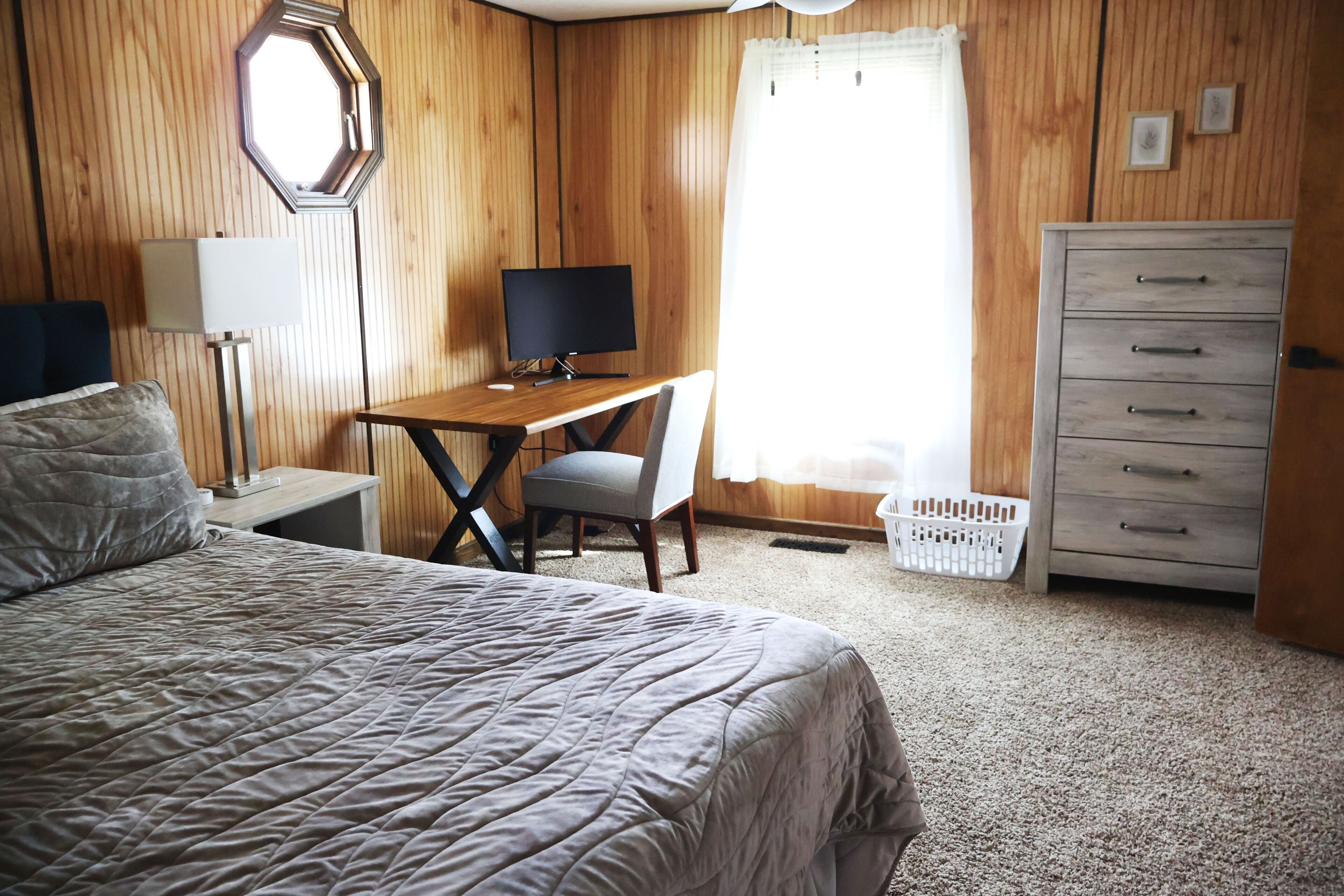 Une chambre à coucher comprenant un lit, un bureau, une chaise, une commode et une fenêtre avec des rideaux.