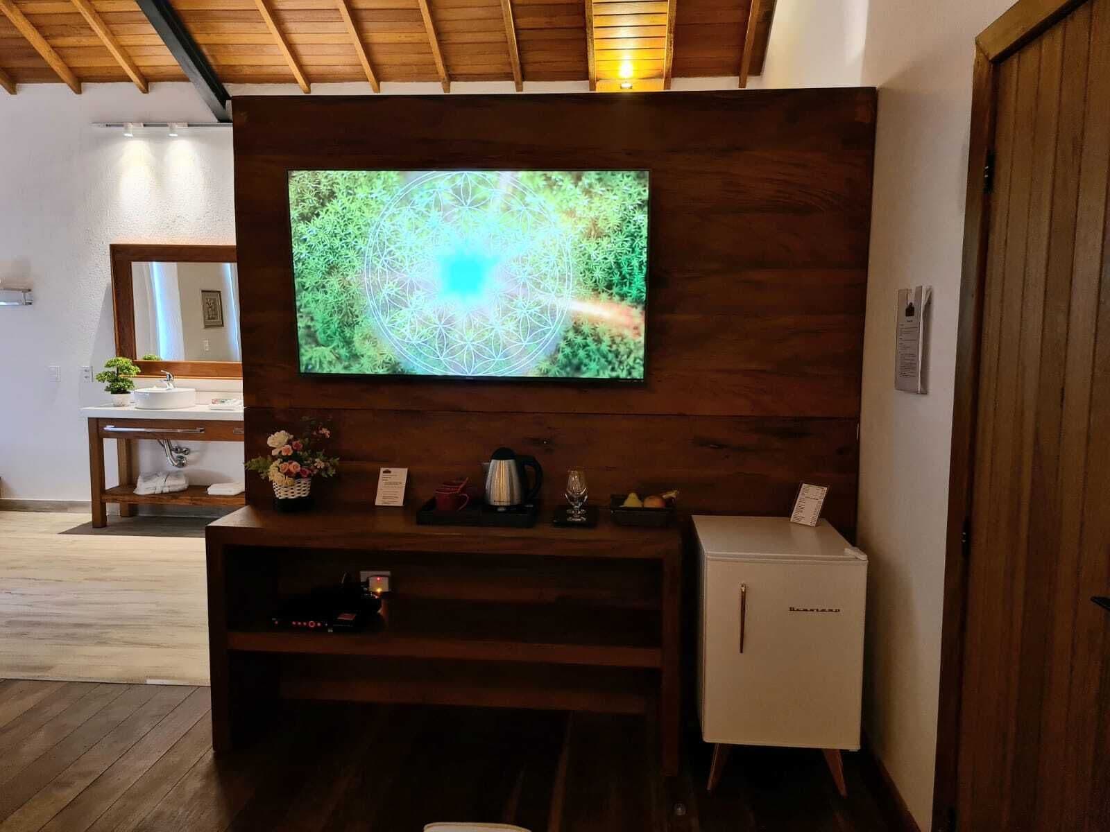 A modern bathroom with a wooden vanity, a mirror, a television mounted on the wall, and a small refrigerator.