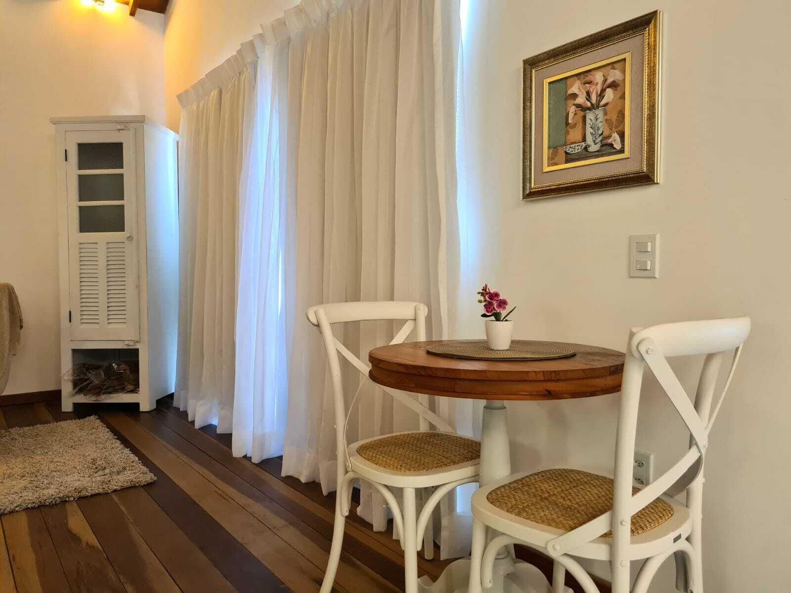 A room with a round wooden table, two white chairs with woven seats, a white cabinet, and a framed picture on the wall.