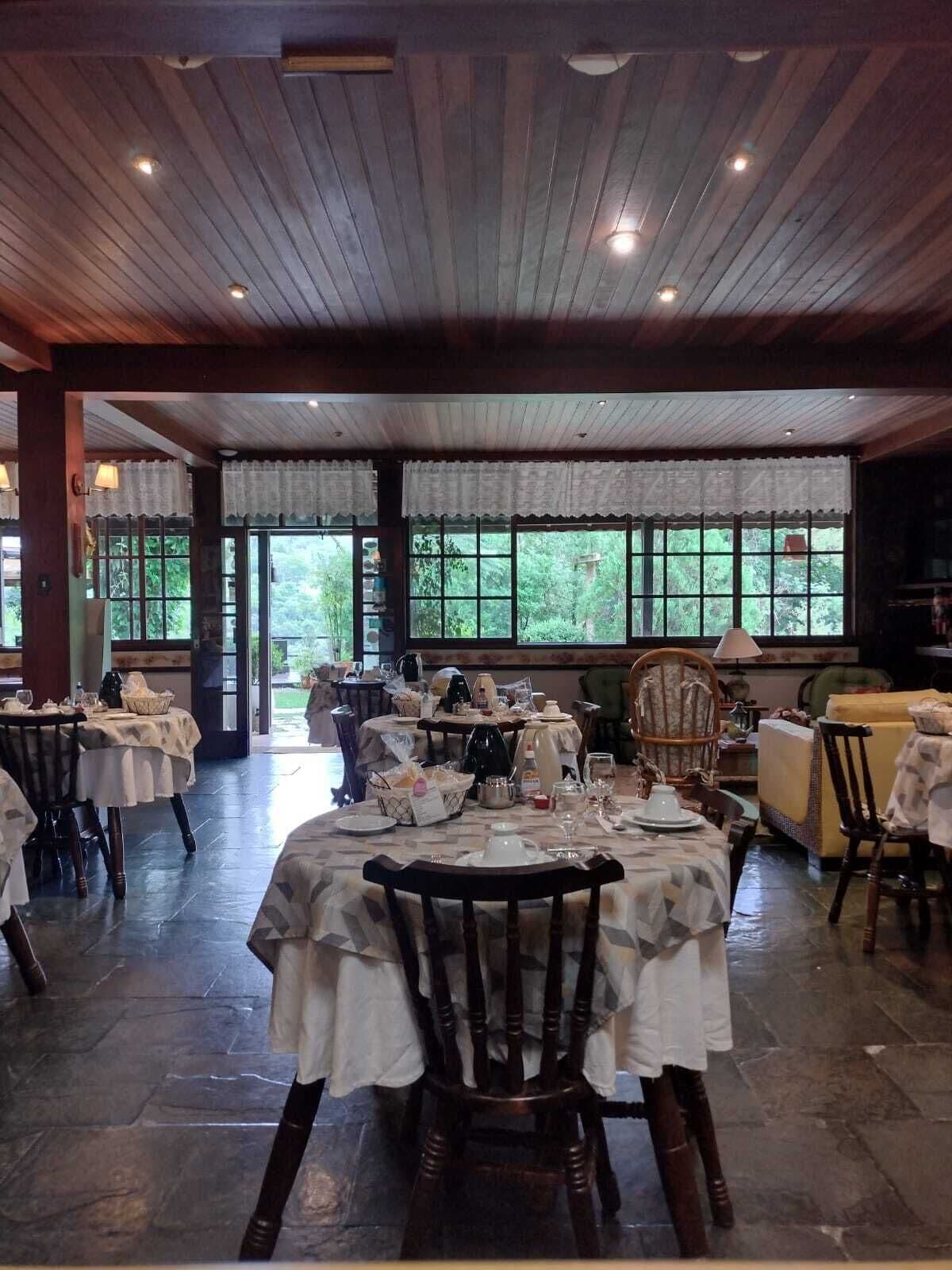 Uma sala de jantar com mesas postas para refeição, janelas amplas com cortinas e teto de madeira.