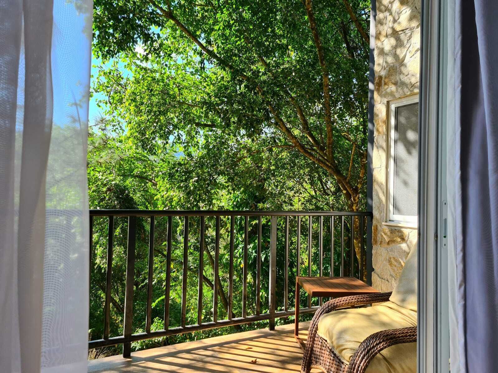 Un balcón con una silla de mimbre, una mesita pequeña y vistas a árboles verdes y frondosos.