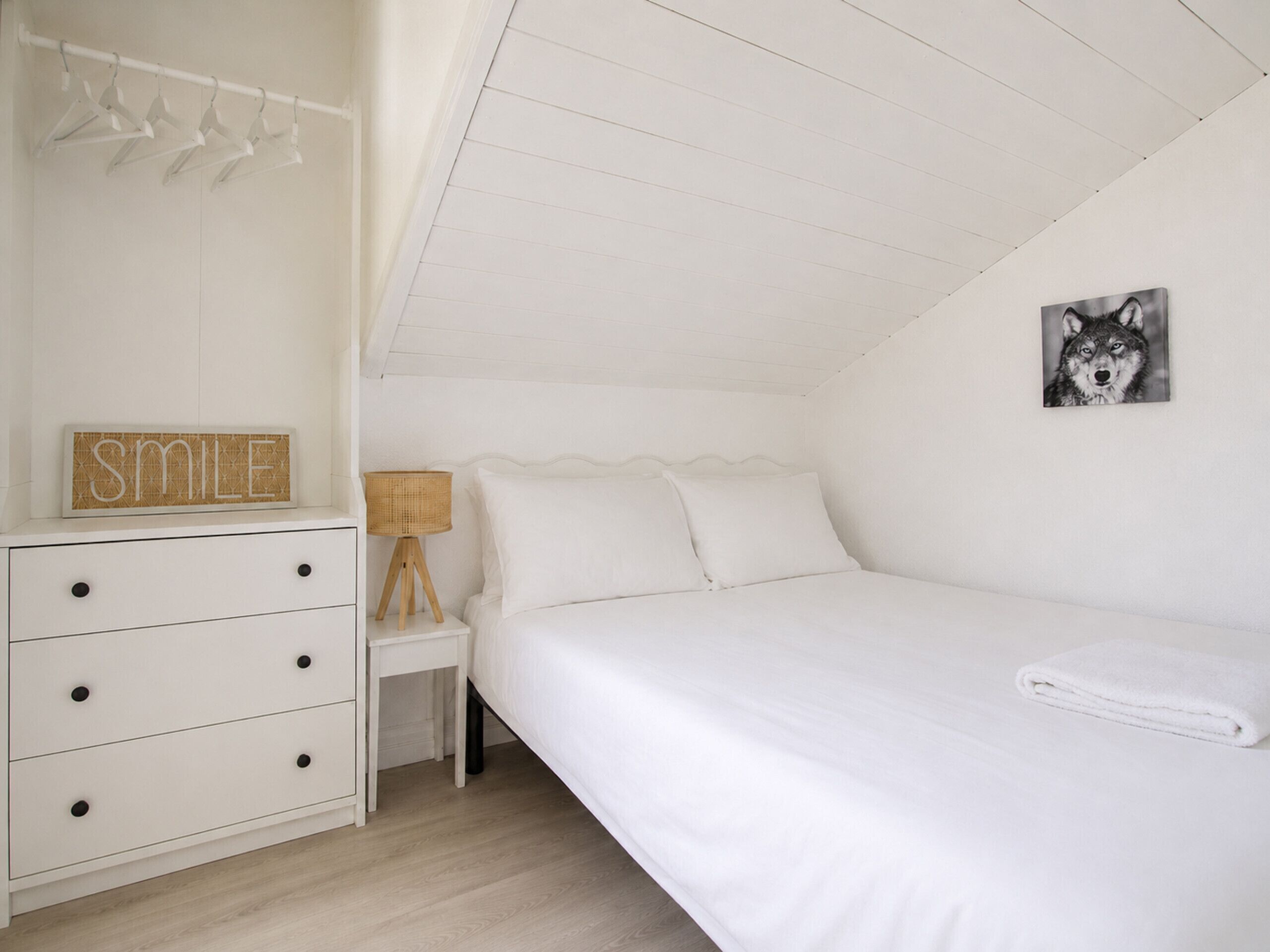 A bedroom with a bed, a dresser, a nightstand, a lamp, a wall-mounted picture, and a 'SMILE' sign.
