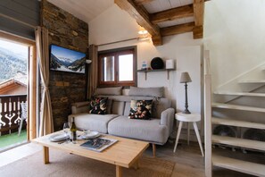 A cozy living room with a sofa, a coffee table, a TV mounted on a stone wall, and a view of the mountains through a window. - Apartment Barberine (Les Houches)