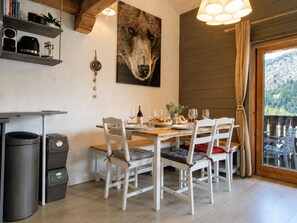 A dining area with a wooden table set for four, white chairs, a large wolf portrait on the wall, and a view through a glass door. - Apartment Barberine (Les Houches)