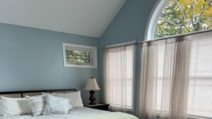 Un dormitorio con una cama, una mesita de noche, una lámpara y una ventana con cortinas traslúcidas.